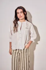 Blusa de algodón peinado color off-white, con cuello mao, cierre frontal de botones, mangas tres cuartos abullonadas con puños con volados, detalles de alforzas y apliques de encaje en el frente y los hombros.