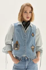 Camisa a rayas verticales de calce recto, con cuello clásico y mangas abullonadas con detalles de frunces y lazos en los puños. Presenta un pequeño bordado en el pecho.