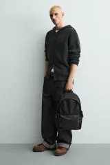 Mochila de tejido técnico en color negro, con compartimento principal con cierre de cremallera, bolsillo frontal con cierre y compartimento interior acolchado para dispositivos electrónicos. Cuenta con parte posterior acolchada, banda de sujeción para equipaje, asa de mano superior y correas de hombro ajustables.