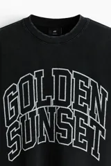 Remera boxy de algodón negro con efecto deslavado, corte oversized y mangas cortas. Presenta un estampado gráfico frontal con la inscripción "Golden Sunset" en estilo universitario.