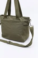 Cartera tipo tote bag de nylon color verde militar, con diseño amplio y liviano. Cuenta con asas superiores reforzadas, correa larga regulable y desmontable, bolsillos frontales y laterales con cierre metálico, y un pequeño aplique con logo en la parte inferior.