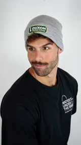 Gorro tipo beanie de punto color gris, con parche rectangular bordado en el frente que incluye el logo de la marca Duxon.