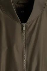 Campera bomber de color verde militar, con cuello elevado acanalado, cierre frontal, bolsillos laterales con cierre y ribetes acanalados en puños y bajo.