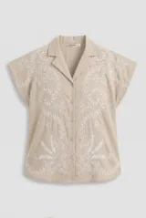 Camisa beige de manga corta y cuello camisero, con bordados florales blancos en el frente y las mangas, y cierre frontal de botones.