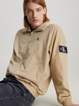 Campera bomber color beige de nailon reciclado, con cuello alto de canalé, puños y bajo elásticos, cierre de cremallera, bolsillos con botón de presión en la cintura, forro de poliéster reciclado e insignia con monograma de Calvin Klein en la manga.