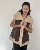 Chaleco estilo gamulán de cuero sintético marrón con interior y bordes de corderito sintético color crema. Presenta cierre frontal con botones a presión y bolsillos laterales.