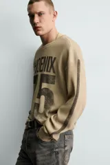 Sudadera de corte relajado confeccionada en tejido con textura waffle, color beige con efecto lavado. Presenta cuello redondo, manga larga con franjas laterales oscuras y estampado frontal con texto y número en contraste.