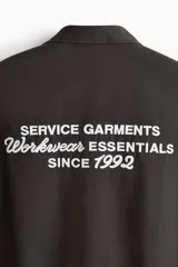 Camisa cargo de manga corta color marrón oscuro, con cuello abierto, botones frontales, bolsillos con solapa en el pecho y el texto 'SERVICE GARMENTS' bordado sobre el bolsillo izquierdo.