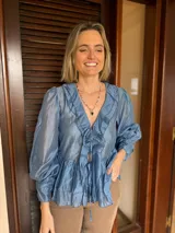 Blusa de tela satinada color azul, con escote en V, volados decorativos en el frente y mangas largas abullonadas con puños ajustados. Presenta un lazo frontal para atar y un diseño con volado en el ruedo.
