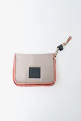 Billetera plegable con cierre de cremallera perimetral. El cuerpo principal es de tela color beige claro, con ribetes y cierre en color salmón/coral. Posee un parche negro con letras blancas en el frente. Incluye un cordón ajustable con tope negro y terminación de cordón en color salmón/coral.