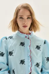 Blusa de algodón en color celeste con estampado floral en negro. Presenta cuello alto con volados y borde en color rojo, mangas abullonadas con puños elásticos tipo smock y cierre frontal con botones.