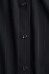 Camisa de manga larga, color negro o azul muy oscuro, con finas rayas verticales en tono más claro. Presenta cuello clásico, cierre frontal de botones y corte holgado con bajo redondeado y aberturas laterales.