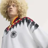 Campera deportiva Adidas de la selección de Alemania, inspirada en el diseño de 1994. Es de color blanco con un estampado geométrico en los hombros que combina los colores de la bandera alemana (negro, rojo y amarillo). Presenta cierre frontal, cuello redondo y el logo de la marca junto al escudo de la DFB en el pecho.
