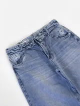 Pantalón de jean estilo wide leg con diseño de cintura cruzada y tiro bajo. Presenta un lavado azul medio con efecto desgastado y un detalle de rotura en la rodilla derecha.