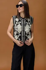 Chaleco tejido sin mangas, color negro con detalles bordados en color crema con diseño de arabescos y flores. Presenta cierre frontal con botones y cuello redondo.