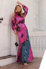 Vestido largo de satén con estampado floral en tonos fucsia y verde azulado. Presenta cuello alto, aberturas en los hombros y mangas largas con puños abotonados.