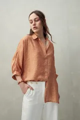 Camisa de manga tres cuartos con cuello clásico y botones frontales. Presenta un estampado sutil en tonos terracota y naranja con textura arrugada.