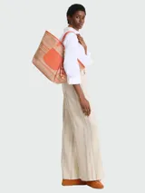 Bolso tipo tote de gran tamaño confeccionado en tejido de paja con rayas multicolores. Presenta un parche frontal de cuero naranja con el logo de la marca grabado, asas de hombro de cuero a juego y un detalle colgante de cuentas de madera. Incluye un cierre de mosquetón metálico en la base.