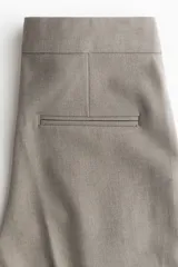Pantalón de vestir color gris topo, tiro alto, con piernas bootcut y raya marcada en el frente y el reverso. Presenta cierre frontal con botón oculto y corchetes, bolsillos al bies y bolsillos traseros ribeteados.