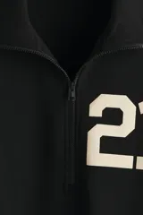 Buzo negro de tejido pesado con interior cepillado suave, cuello elevado con medio cierre y hombros ligeramente caídos. Presenta un estampado frontal con el número "21" en color beige. Puños y bajo en punto acanalado.