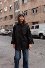 Parka liviana color negro con capucha, cierre frontal oculto y bolsillos delanteros con tapa. Presenta mangas tres cuartos con detalle de botones y ajuste en la cintura.