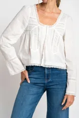 Blusa blanca de algodón con escote en U y mangas largas acampanadas. Presenta detalles de encaje en el escote, el frente y los puños.