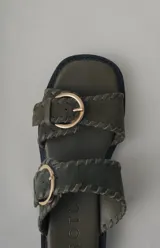 Sandalia de plataforma color verde militar con doble tira ancha. Las tiras tienen un acabado de gamuza con pespuntes decorativos y hebillas metálicas doradas. La suela es gruesa y negra.