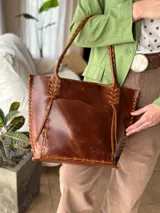 Cartera tipo shopper de cuero genuino color marrón con estampado texturado tipo cocodrilo. Presenta detalles de costuras y tiras trenzadas artesanales en los bordes y en las asas de hombro.