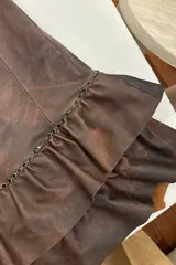 Vestido strapless corto confeccionado en cuero ovino color óxido. Presenta un diseño ajustado en el torso con costuras verticales y una falda con doble volado. Incluye detalles de pequeñas tachas metálicas oscuras en la unión de la falda.