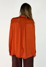 Camisa de satén color naranja óxido, con cuello mao y lazo para anudar. Presenta volados decorativos en el frente y en las mangas, las cuales son largas y terminan en puños abotonados.