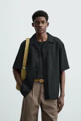 Camisa de lino color negro con calce relajado, cuello bowling y manga corta. Presenta cierre frontal con botones a tono y aberturas laterales en el bajo.