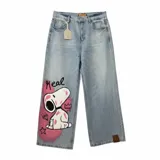 Jean de corte wide leg en denim azul claro con lavado desgastado. Presenta una ilustración pintada a mano de Snoopy en la pierna derecha, acompañada por la palabra 'Real', una corona y una estrella. Incluye etiqueta de cuero en la cintura y en el ruedo.