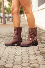 Bota corta de cuero color marrón, con taco cuadrado y punta fina. Presenta una correa con tachas pequeñas y una argolla metálica decorativa alrededor del tobillo. Cuenta con cierre lateral.