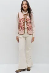 Chaleco de lana color beige con bordados florales artesanales en tonos marrones y blancos. Presenta un diseño sin mangas con volados en los hombros y cierre frontal mediante lazos negros ajustables.