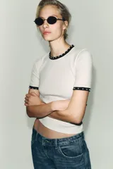 Camiseta blanca de manga corta y cuello redondo, con ribete negro decorado con perlas blancas en el cuello y los puños.