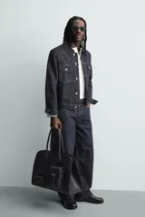 Bolso de viaje negro con acabado grabado y diseño de múltiples bolsillos frontales. Dispone de un compartimento principal con cierre magnético, un bolsillo interno con cremallera y tres bolsillos exteriores de diversos tamaños con cierre de imán. Incluye dos asas de mano superiores.