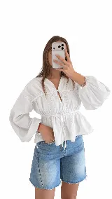 Blusa blanca de manga larga amplia, con escote en V y frunces en el cuerpo. Posee cordones ajustables con lazos en el frente y en la cintura.