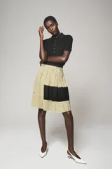 Falda de tiro medio confeccionada en hilatura de lino, color beige con bloques negros y detalles de abalorios y ojales metálicos aplicados a contraste. Cuenta con cierre lateral con cremallera oculta en costura.