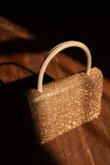 Cartera de mano color dorado, con diseño rectangular y superficie completamente cubierta de brillos. Cuenta con un asa superior rígida.