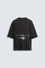 Camiseta de punto con corte boxy fit, cuello redondo y manga corta. Color negro con efecto lavado y terminaciones irregulares. Presenta un estampado frontal con texto y un gráfico circular en tonos blanco y rojo.