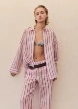 Camisa de corte oversize confeccionada en tejido de ramio, con diseño de rayas verticales en tonos rosados y oscuros. Presenta cuello camisero clásico, cierre frontal con botones y mangas largas con puños.