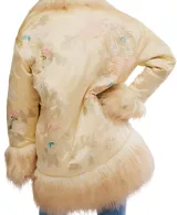 Chaqueta estilo kimono o bata, color crema o amarillo pálido, con acabado satinado y bordados florales sutiles. Presenta un ribete y puños voluminosos de pelo sintético largo y rizado en tono beige/crema.