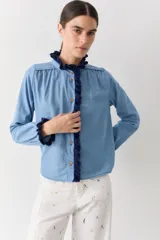 Camisa de algodón estilo denim con corte regular, cuello redondo y cierre frontal con botones. Presenta detalles de volados tejidos a mano en crochet en color azul marino con bordes rojos en el cuello y la cartera frontal.