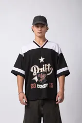 Camiseta de estilo deportivo inspirada en el fútbol americano, con diseño de bloques de color en negro y blanco. Presenta cuello en V con detalles a rayas, mangas cortas anchas y estampado frontal con efecto desgastado que incluye gráficos y texto.