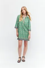 Túnica verde con estampa geométrica de cuadrados en verde más oscuro y blanco. Tiene escote en V con ribete marrón y mangas tres cuartos con ribete geométrico en puños y ruedo.