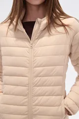 Campera acolchada tipo puffer, color beige claro, con capucha y cierre frontal. Presenta costuras horizontales y un calce entallado.