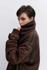 Cazadora de estilo bomber con diseño híbrido, combinando paneles de tela técnica en las mangas con un cuerpo de efecto piel desgastada y forro interior de corderito sintético. Presenta cierre frontal, cuello amplio y puños elásticos.