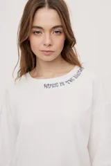 Remera de manga corta y cuello redondo, de corte oversized en color marrón, con texto bordado o estampado a lo largo del escote en el hombro izquierdo.