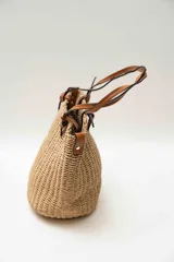 Bolso de mano tipo tote confeccionado en tejido texturizado efecto rafia en color beige, con asas dobles finas de cuero sintético marrón y detalles de nudos en los extremos.