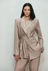 Blazer de sastrería color beige con textura satinada, solapas de muesca, bolsillos con solapa y detalle fruncido con cordones ajustables en la cintura.